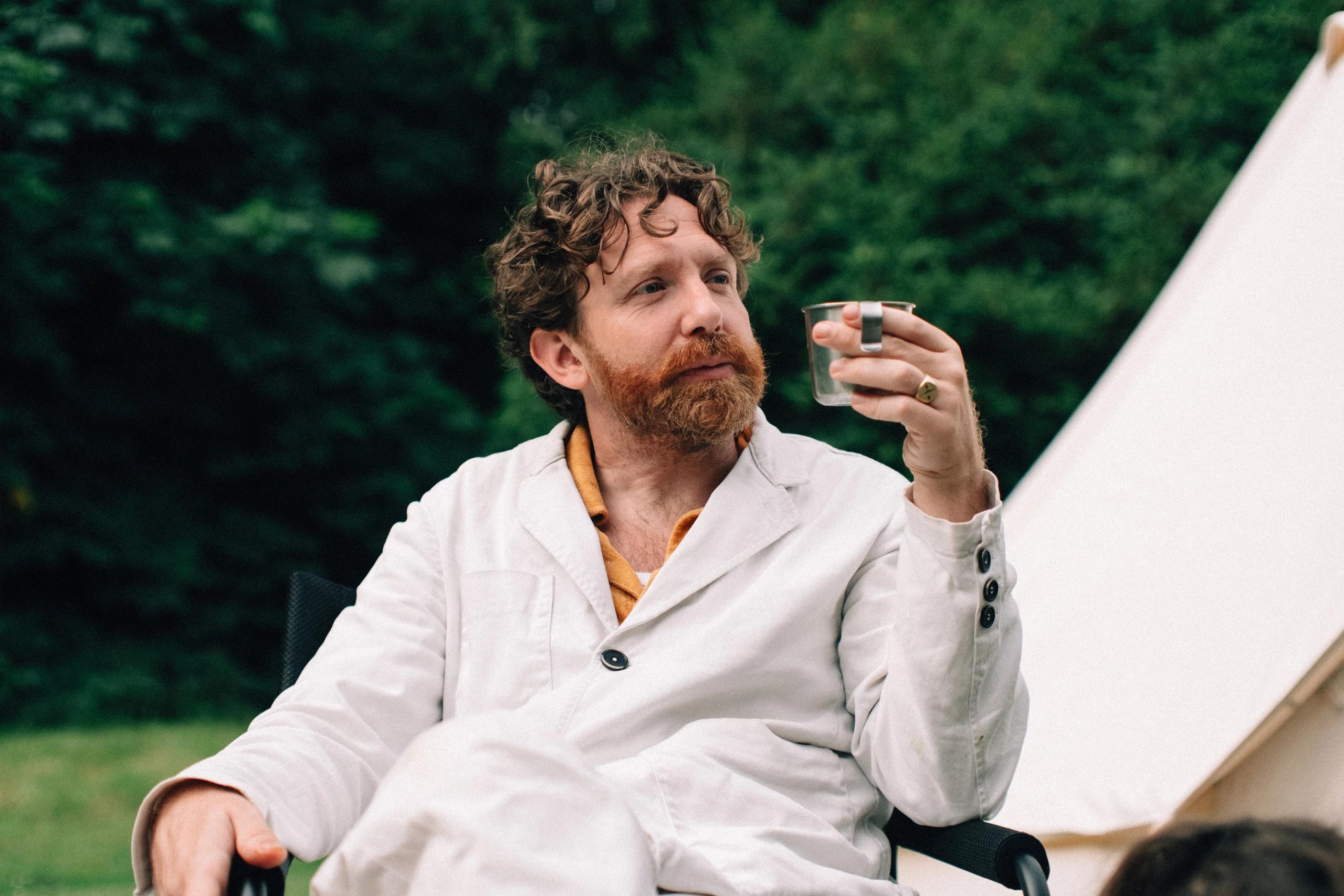 Bearded man examining a small glass beside a tent and green trees