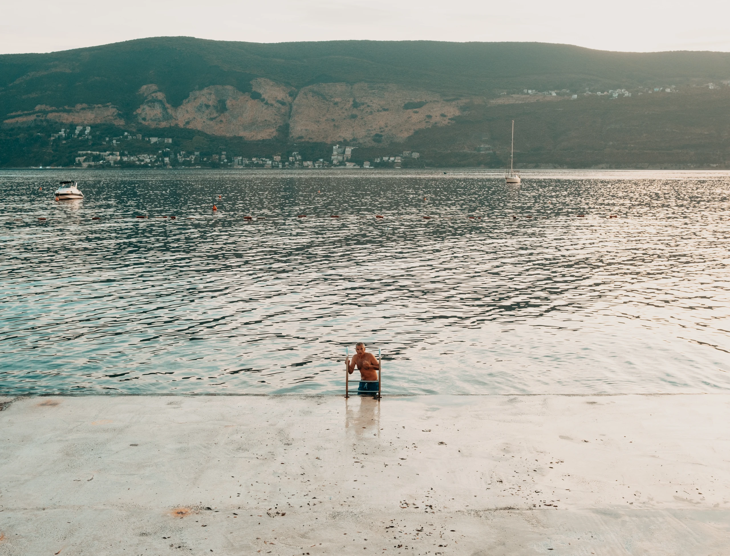 Lone swimmer at a waterfront ladder overlooking a wide bay and distant hills