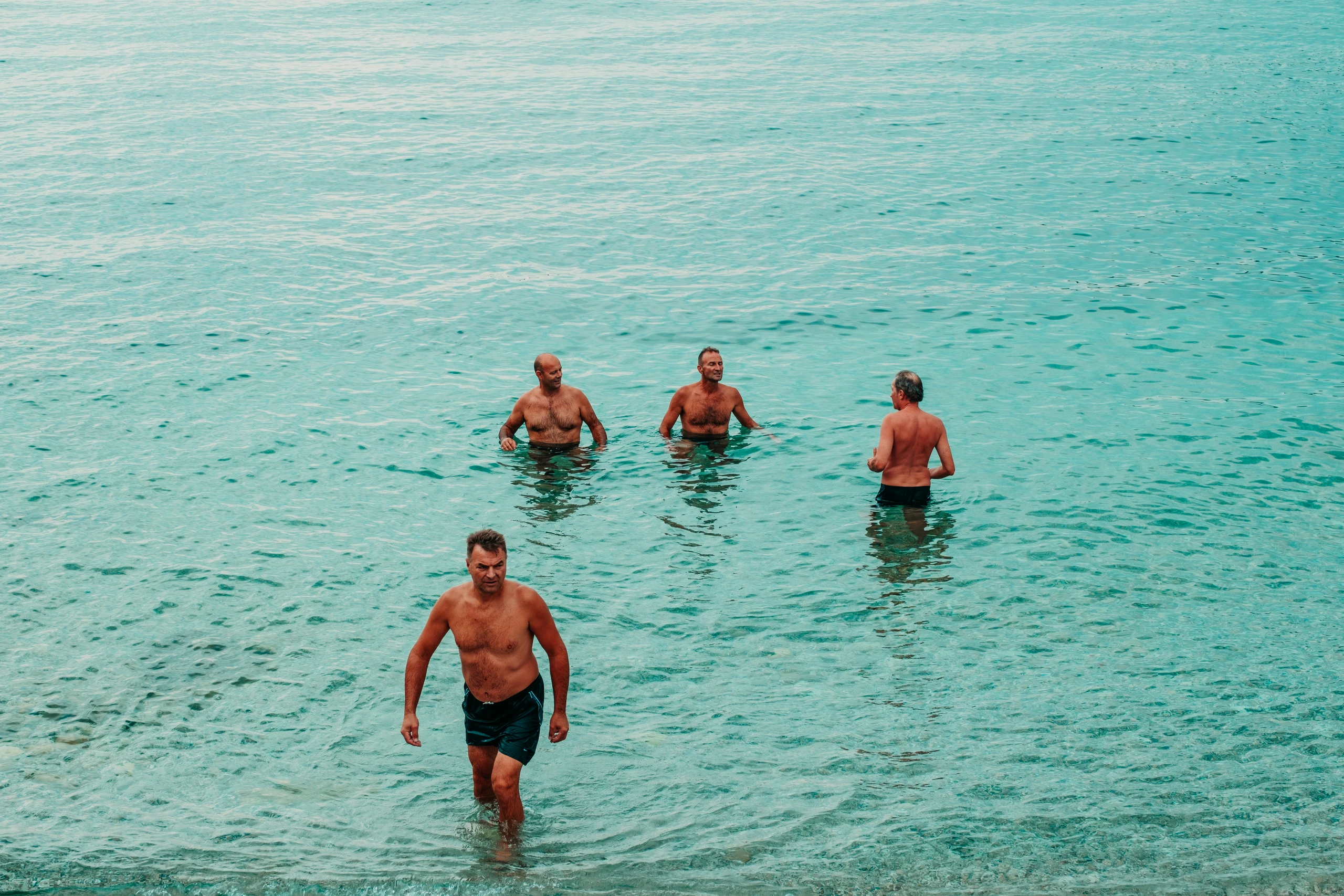 Four men standing in bright turquoise water beside a waterfront