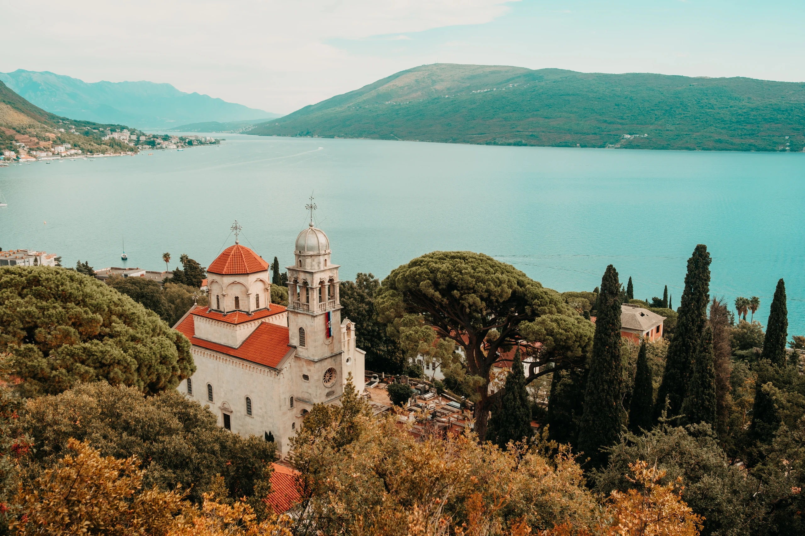 Church with red roofs and cypress trees overlooking a blue mountain bay