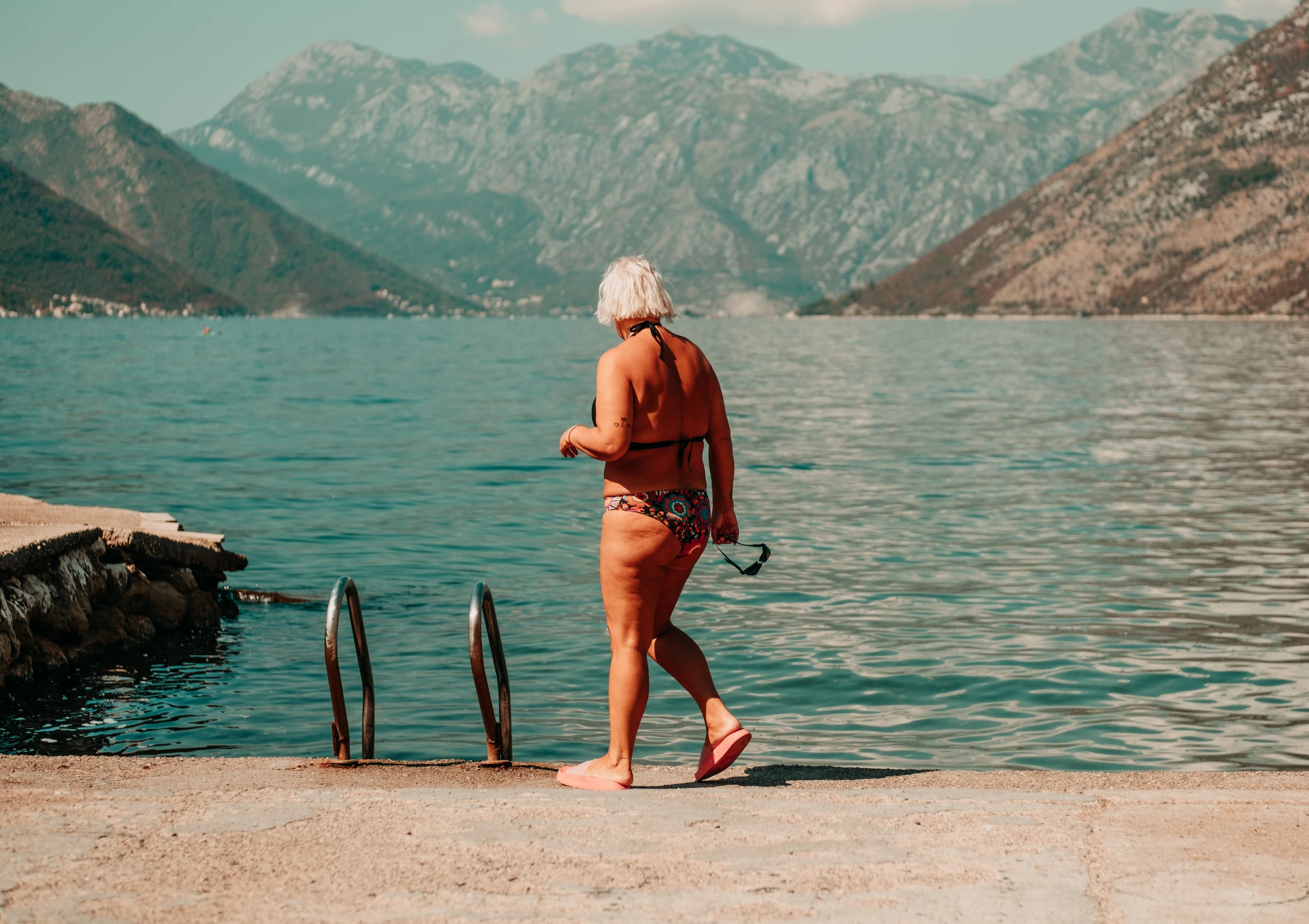 Woman in a swimsuit walking towards a swimming ladder with mountains across the bay