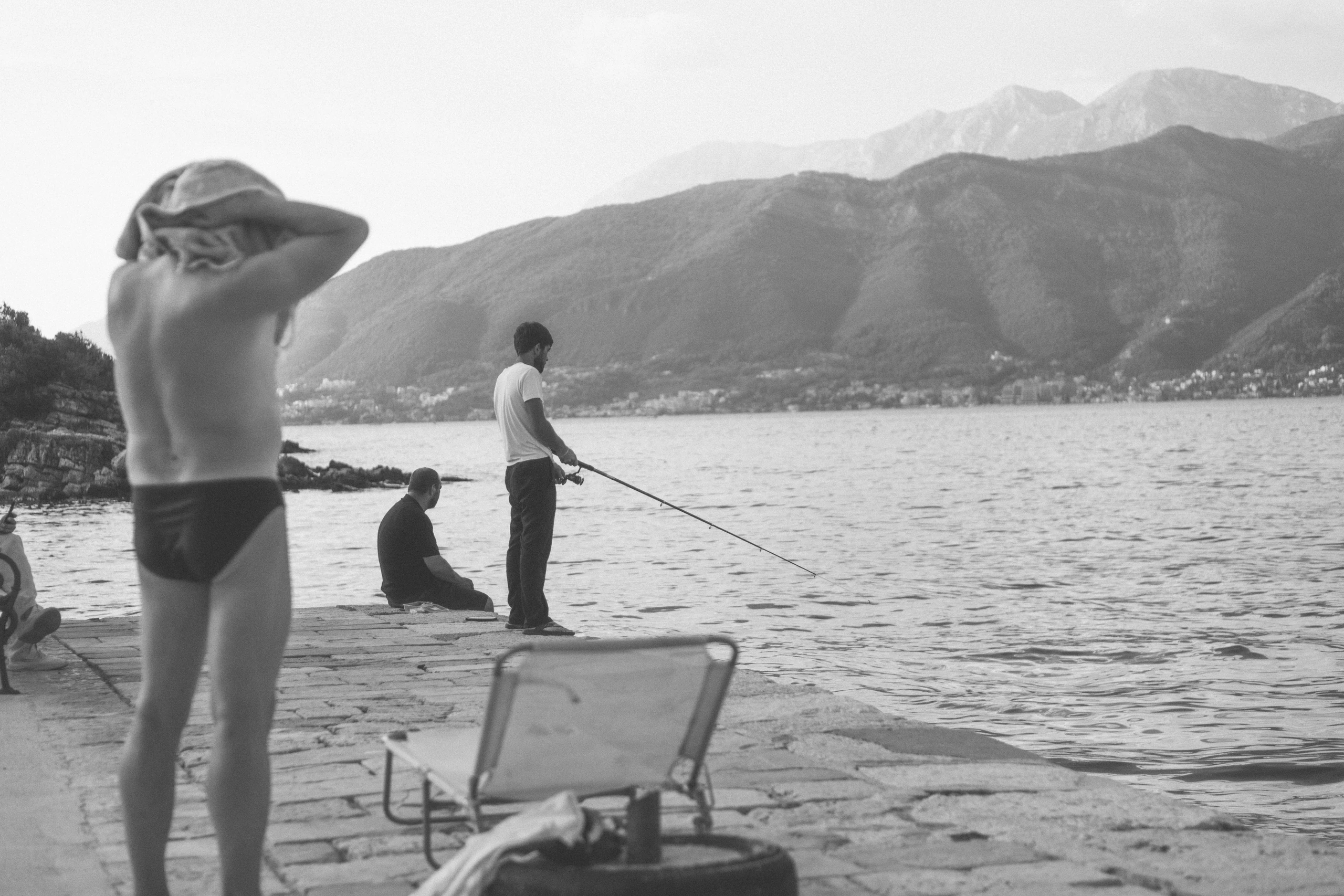 Black-and-white scene of swimmers and a person fishing beside a mountain bay