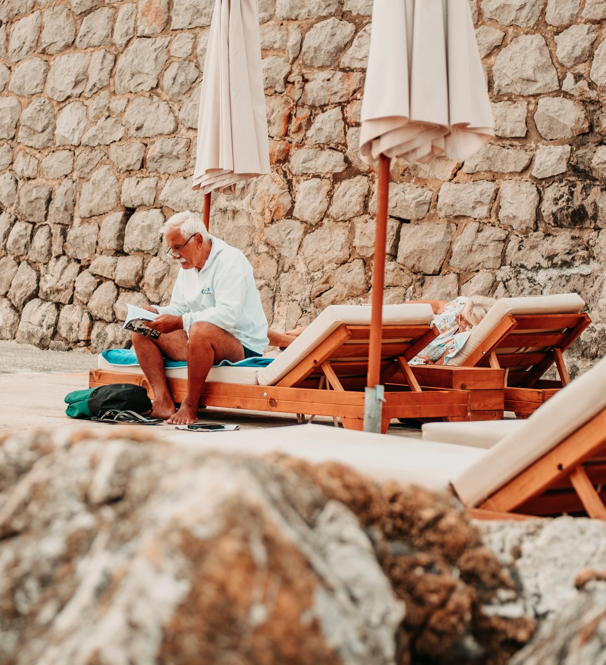 Older man reading beside wooden sun loungers and a stone wall